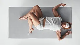 Top view: A person in pyjamas and a sleep mask is lying on their back on a mattress, with their legs bent to the side and their arms stretched out to the side.