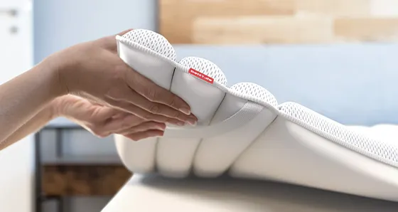 Photo: Two hands hold the BODYGUARD topper (100x220 cm) slightly above the mattress, so that the fastening loop and the contouring of the topper are clearly visible.