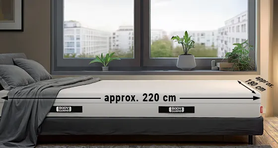 A BODYGUARD® mattress is lying in a bedroom. There is an arrow labeled “approx. 220 cm” on the long side and an arrow labeled “approx. 180 cm” on the wide side.