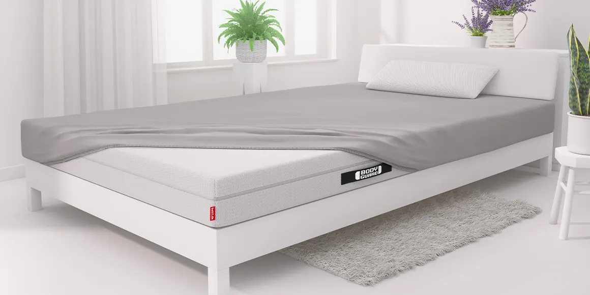 Photo: In a bright white bedroom stands a large white bed with a BODYGUARD mattress, covered with a fitted sheet on only three corners.