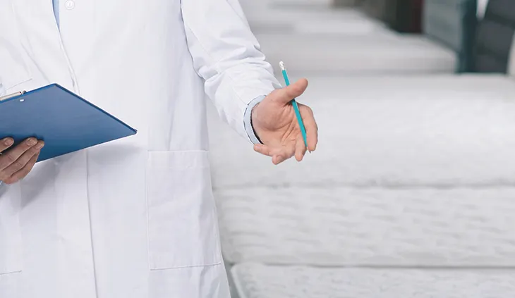 Eine Person in weißem Kittel hält ein Klemmbrett und einen Kugelschreiber in der Hand. Daneben verschiedene Matratzen.