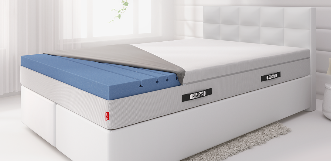Photo: A bed with a BODYGUARD Box Spring Mattress 180x220 cm in a bright bedroom. The mattress cover is folded back halfway, revealing the upper section of the two-part mattress core