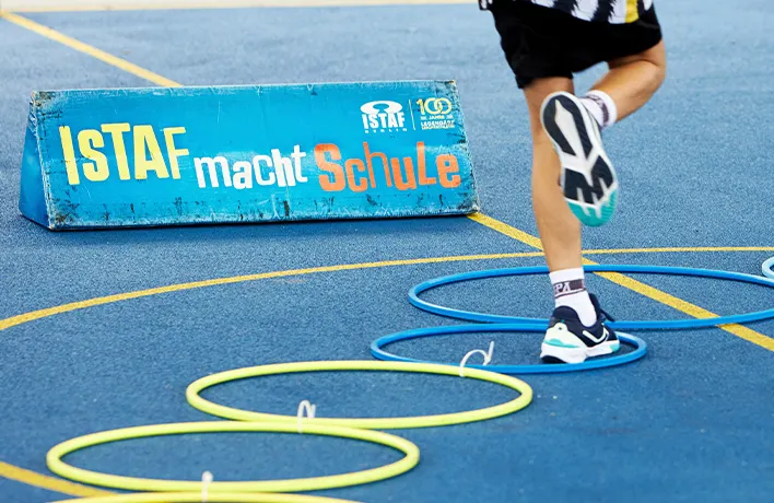 Auf einem Spielfeld liegt ein Parkour aus Hoola-Hoop-Reifen und Kinderbeine springen hindurch, dahinter ein Banner mit Text: ISTAF macht Schule.