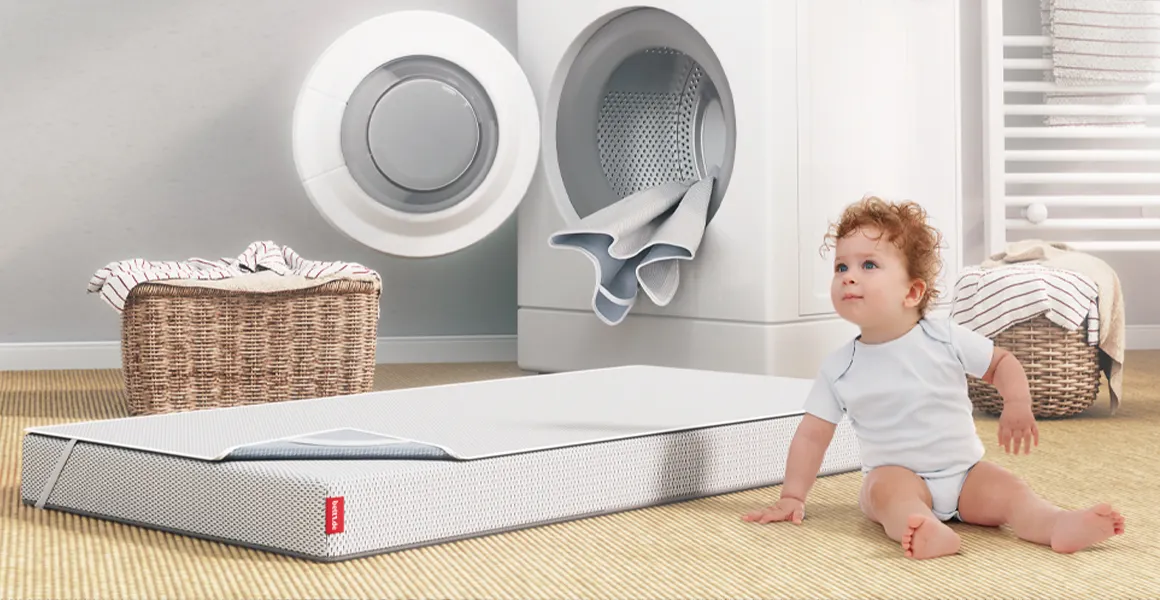 In einem Badezimmer sitzt ein Kleinkind vor einer Waschmaschine. Daneben liegt eine SuperBreeze Kindermatratze auf dem Boden. Sie ist mit dem BODYGUARD Nässeschutz bezogen.