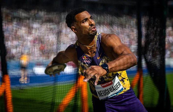 A track and field athlete, with his upper body turned as if to throw, stands in front of a safety net.