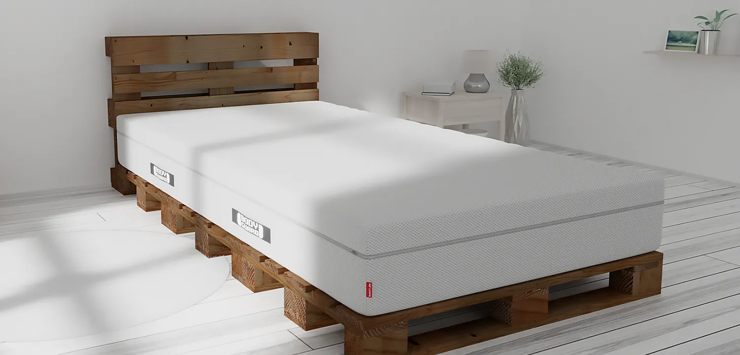 In a bright bedroom stands a dark wooden pallet bed, topped with the BODYGUARD box spring mattress, 100x220 cm.