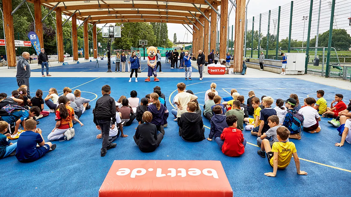 Zahlreiche Kinder sitzen auf einem Sportplatz und blicken in Richtung des Maskottchens vom bett1.de Multisportevent.