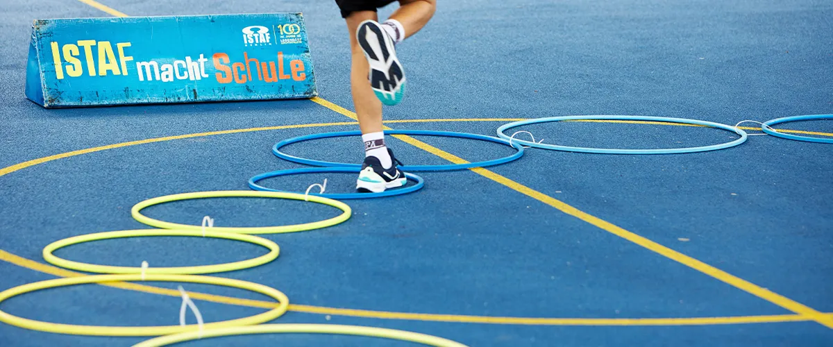Auf einem Spielfeld liegt ein Parkour aus Hoola-Hoop-Reifen und Kinderbeine springen hindurch, dahinter ein Banner mit Text: ISTAF macht Schule.