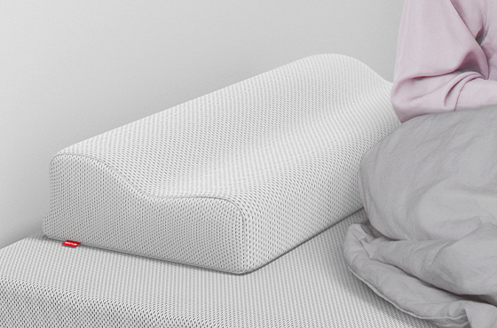 The BODYGUARD neck support pillow is lying on a BODYGUARD mattress, next to it is a person sitting halfway under a duvet.