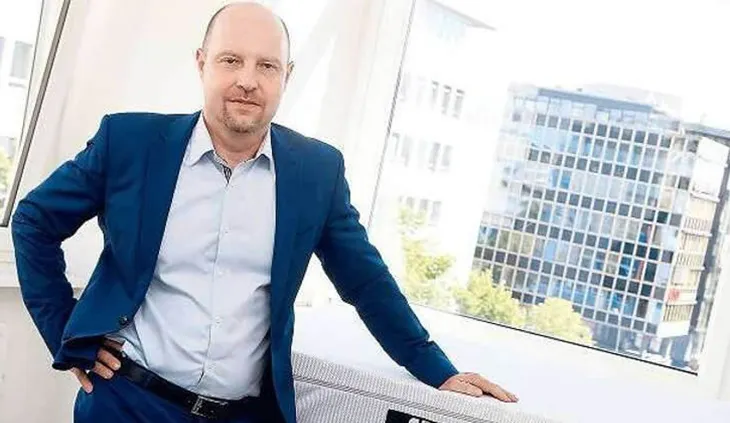bett1-Gründer Adam Szpyt lehnt an einer BODYGUARD Matratze. Im Hintergrund steht ein Gebäude mit vielen Fenstern.
