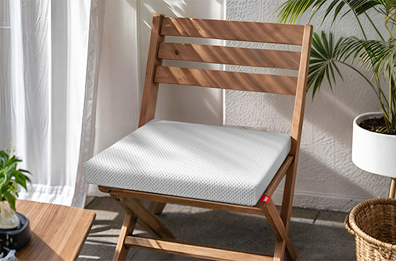 A wooden folding chair stands in the sun. The BODYGUARD® seat cushion lies on the seat. Next to it are plants. 