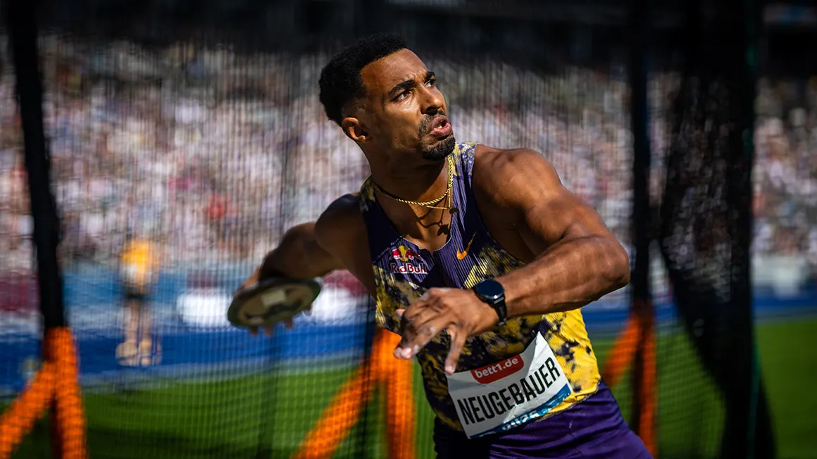 A track and field athlete, with his upper body turned as if to throw, stands in front of a safety net.
