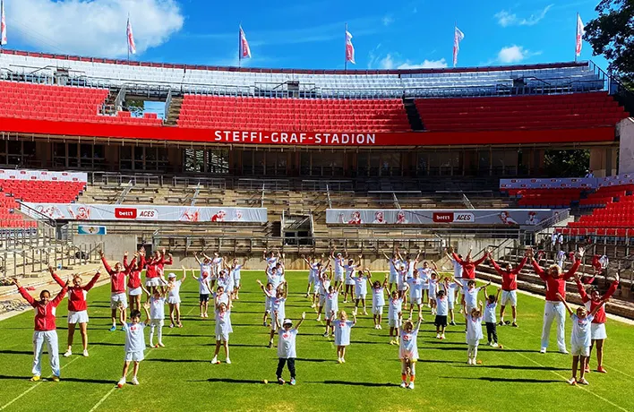 Vor einer roten Tribüne stehen Kinder und Erwachsene in roter und weißer Sportbekleidung in Reihen und heben beide Arme in die Luft.