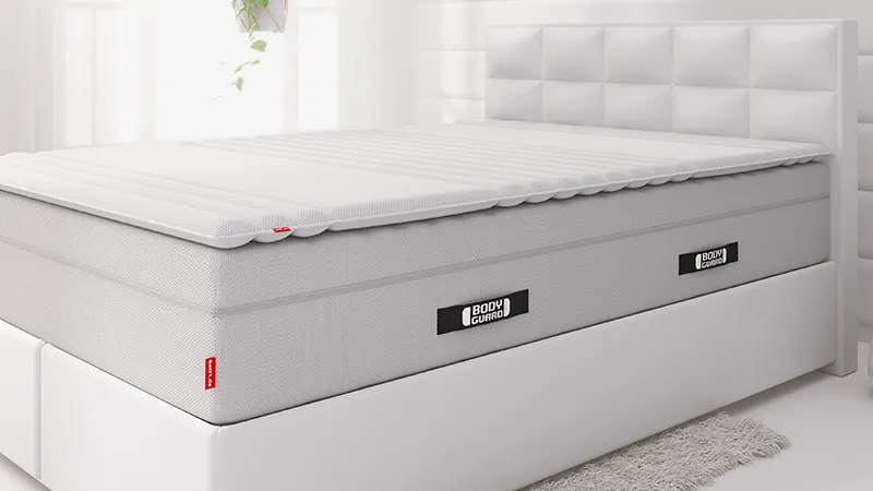 The extra-high BODYGUARD mattress lies on a white box spring bed, with the 140x200 cm topper on top.