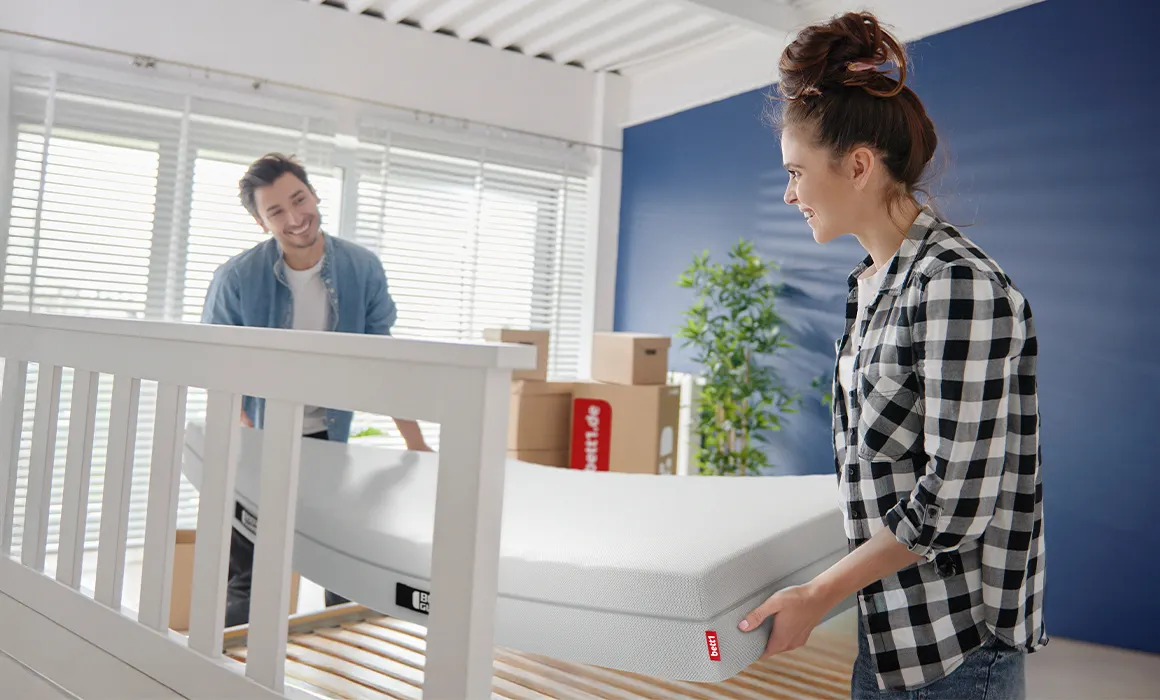 Two people lifting a mattress onto a slatted frame.