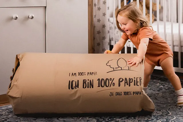 Photo: In front of a crib lies a children's mattress rolled up in paper with the inscription: “I am 100% paper”.  A toddler stands next to it.