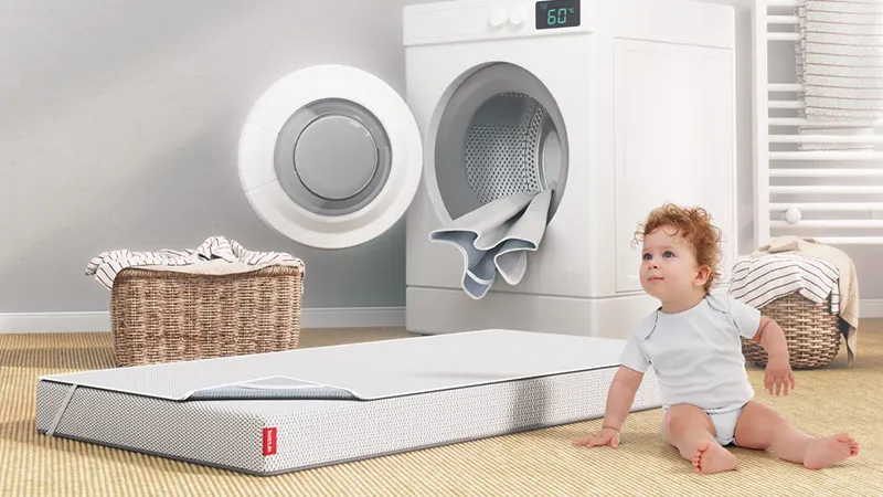 The SuperBreeze® Kids Mattress covered with a wetness protection pad lies in front of an open washing machine from which another wetness protection pad is hanging.