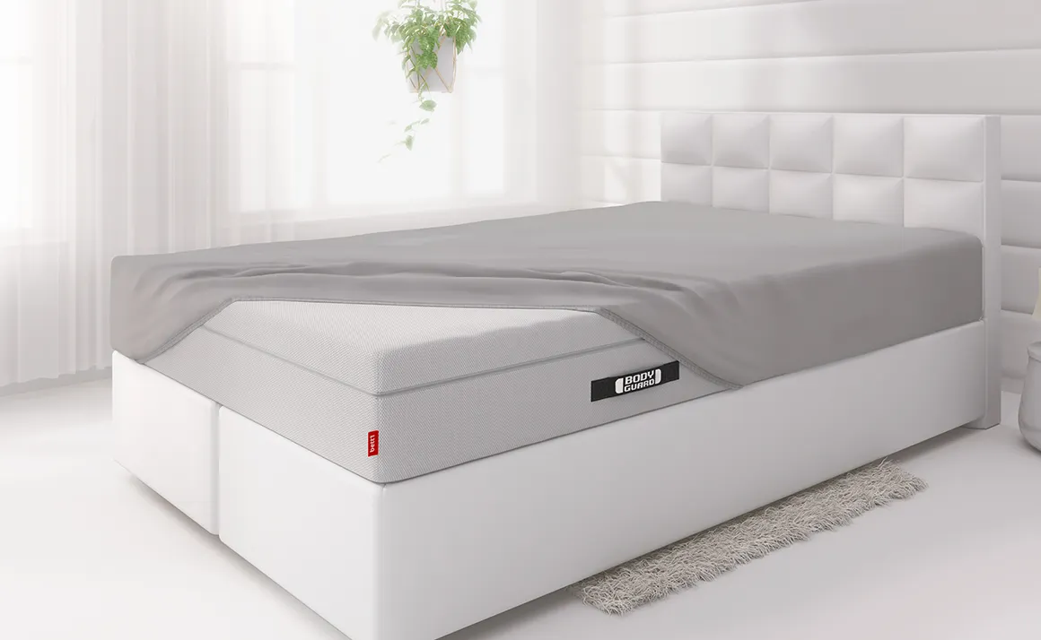  The BODYGUARD® Boxspring mattress is covered with a fitted sheet except for one corner and lies on a boxspring bed in a light-coloured bedroom	