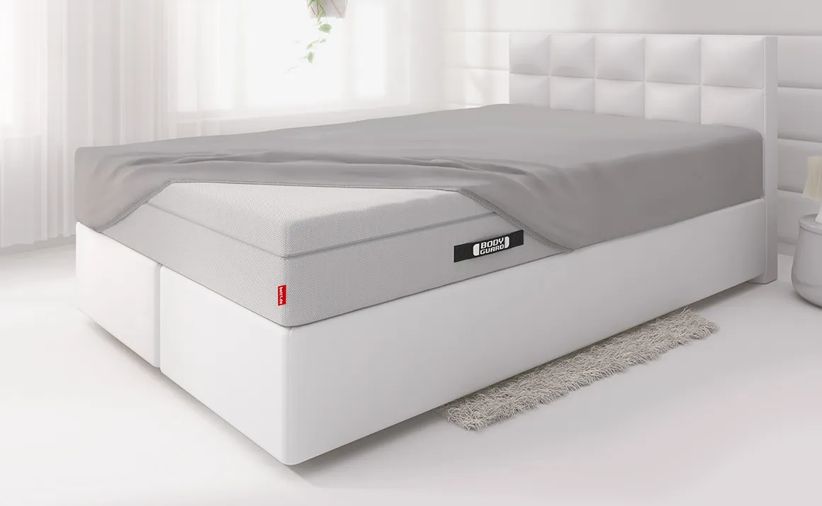  The BODYGUARD® Boxspring mattress is covered with a fitted sheet except for one corner and lies on a boxspring bed in a light-coloured bedroom	