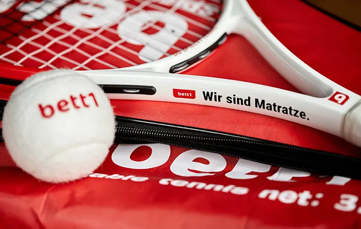 Foto: Im Fokus ein Tennisschläger mit der Aufschrift „bett1 – Wir sind Matratze“; davor liegt ein Tennisball mit der Aufschrift „bett1“. 