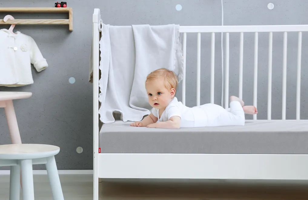  A baby lies in a cot on a baby mattress, which is covered with the children's fitted sheet in silver.	