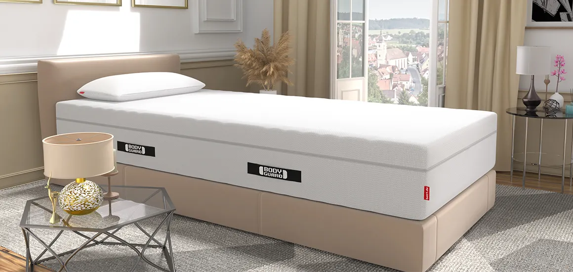The BODYGUARD mattress on a bed in a bright bedroom. The BODYGUARD Visco pillow is on the mattress. In the background, floor-to-ceiling windows are open, offering a view of a city; in the foreground, there is a side table with a lamp.