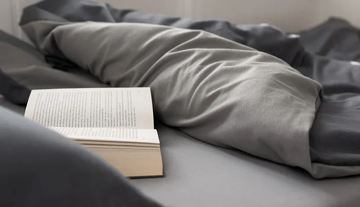  Foto: Unter einer zurückgeklappten Bettdecke liegt ein aufgeschlagenes Buch auf einem Laken.
