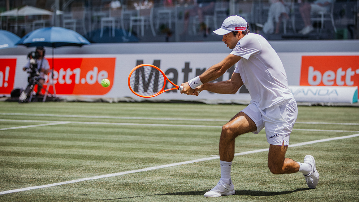 Auf einem Tennisplatz: Person in heller Kleidung und Kappe schlägt auf einem Rasenplatz einen tiefen Ball. Im Hintergrund sind bett1.de-Werbebanden zu sehen.