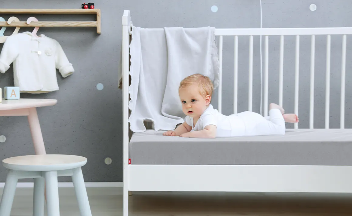 A baby lies in a cot on a baby mattress, which is covered with the children's fitted sheet in silver.