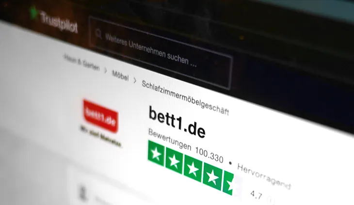Auf einem Bildschirm ist die Trustpilot-Website geöffnet. Zu sehen ist das bett1-Logo. Daneben 4,7 von 5 Sternen. Text: Bewertungen 100.330, hervorragend