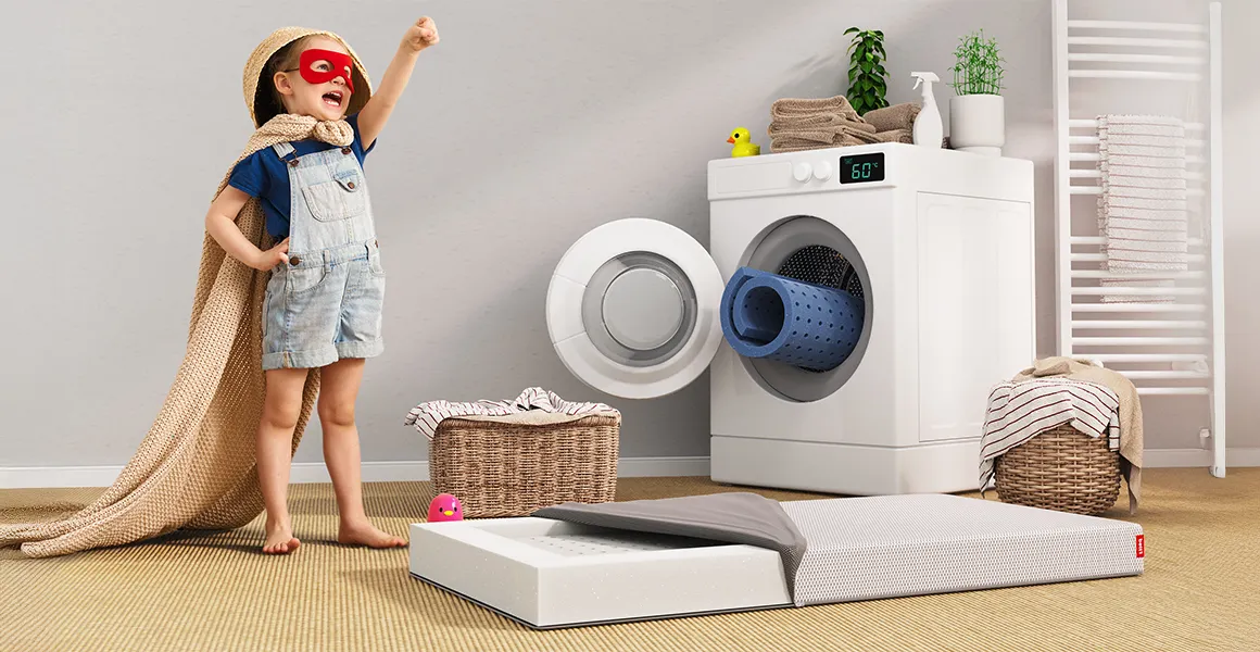The BODYGUARD children's mattress lies on the floor next to a child in a superhero costume. The mattress cover is open, the removable lying surface has been removed and is rolled up in a washing machine behind the mattress.