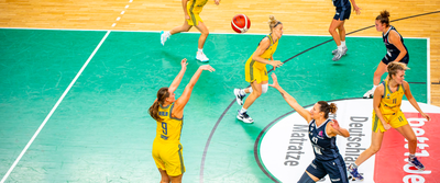 Photo from behind a basketball hoop in a sports hall. People in yellow and blue jerseys are standing on the court.