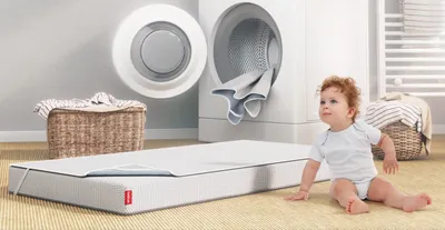A children’s mattress with BODYGUARD Kids Mattress Protector is sitting in front of an open washing machine. Next to it there is sitting a toddler, looking on curiously.