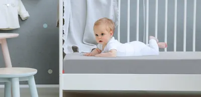 A baby is lying on their tummy in a cot. The cot is fitted with the SuperBreeze Kids Mattress and the BODYGUARD Fitted Sheet 70x140 for children.