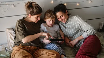 Photo: Two people and a child lying in bed, looking together at a tablet the child is holding.