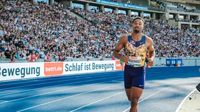 A track and field athlete runs on a tartan track in front of packed stadium stands.