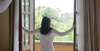 Photo: A person stands at a wide-open balcony door, arms on either side of the double doors, looking out at the greenery.