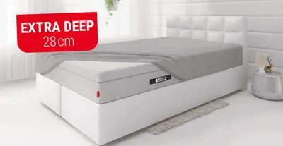 The BODYGUARD box spring mattress lies in a bed and is covered with the extra-high BODYGUARD fitted sheet, except for one corner. A red box with text: extra deep 50 cm. 