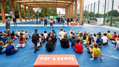 A large number of children are sitting on a sports pitch, looking towards the mascot of the bett1.de multi-sport event.