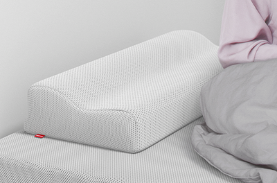 The BODYGUARD neck support pillow is lying on a BODYGUARD mattress, next to it is a person sitting halfway under a duvet.