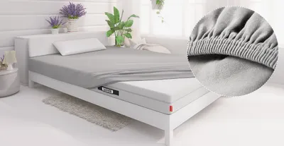 In einem Schlafzimmer liegt eine BODYGUARD Matratze in einem Bett, das mit dem Spannbettlaken 140x200 cm bezogen ist. Ein kreisförmiger Ausschnitt zeigt den Spanngummi in Nahaufnahme.