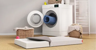 The SuperBreeze Kids Mattress with its cover half open is in front of a washing machine. A washable mattress cover is rolled up and sticking out of the washing machine.