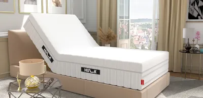 The HULK with the headboard raised on a bed in a bedroom. There is an open window in the background. In the foreground is a side table with a lamp on it.