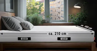 Eine BODYGUARD® Matratze liegt in einem Schlafzimmer. An der Längsseite ist ein Pfeil mit "ca. 210 cm", an der Breitseite ein Pfeil mit "ca. 100 cm" beschriftet.