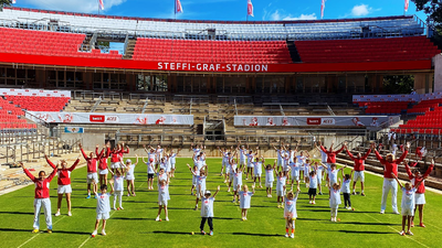 In front of a red grandstand, children and adults dressed in red and white sportswear are standing in rows, raising both arms into the air.