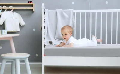 Ein Baby liegt in einem Kinderbett auf einer Babymatratze, die mit dem Kinder-Spannbettlaken in Silber bezogen ist.	